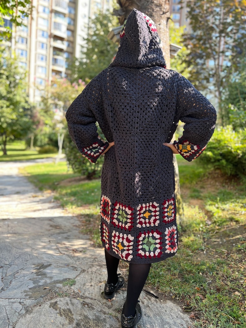 May include: A long, dark gray crocheted cardigan with a hood. The cardigan has a colorful geometric pattern at the bottom hem.