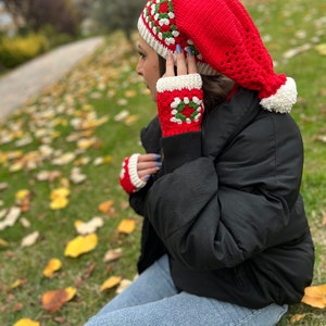 May include: A red crocheted hat with a white pom-pom and a white and green geometric pattern. The hat is worn by a person wearing a black jacket and blue jeans.
