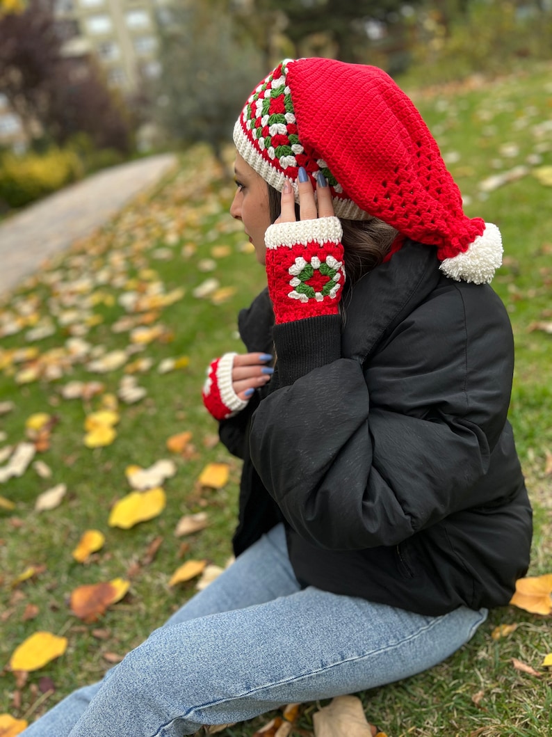 May include: A red crocheted hat with a white pom-pom and a white and green geometric pattern. The hat is worn by a person wearing a black jacket and blue jeans.