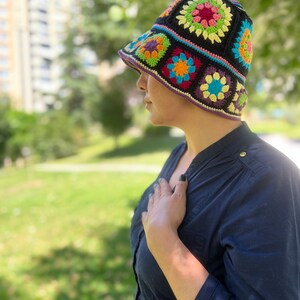 May include: A black crochet bucket hat featuring colourful granny square designs. The hat has a wide brim and is crafted using a range of colours, including red, orange, yellow, green, blue, and purple.
