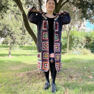 May include: A long, gray crochet cardigan with a hood. The cardigan is made with colorful granny squares and has pockets. The cardigan is being worn by a person standing in a grassy area.