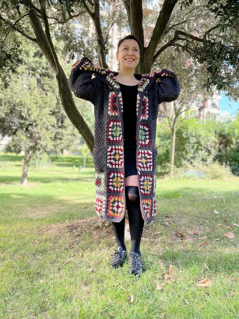 May include: A long, gray crochet cardigan with a hood. The cardigan is made with colorful granny squares and has pockets. The cardigan is being worn by a person standing in a grassy area.