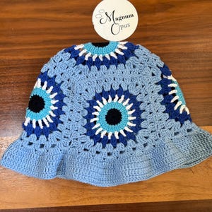 Può includere: Cappello a secchiello in crochet blu con un designo di occhio turco bianco e blu. Il cappello ha un'ampia tesa ed è realizzato in un materiale morbido e leggero.
