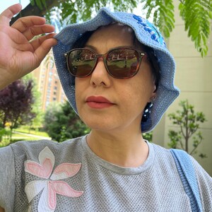 May include: A woman wearing a light blue crocheted sun hat with a floral design, brown sunglasses, and a grey t-shirt with a pink and white flower applique.