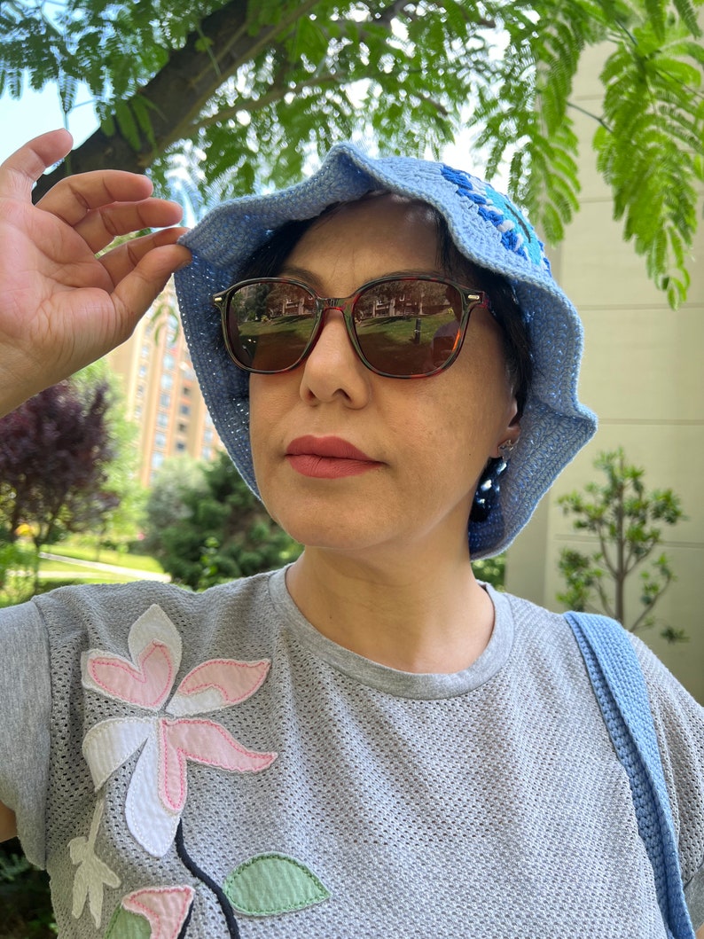 May include: A woman wearing a light blue crocheted sun hat with a floral design, brown sunglasses, and a grey t-shirt with a pink and white flower applique.