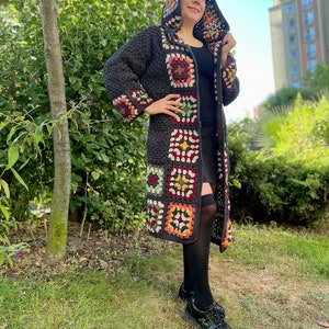 May include: A long, gray crochet cardigan with a hood. The cardigan has colorful granny square patches on the front and sleeves. The model is wearing black tights and black shoes.