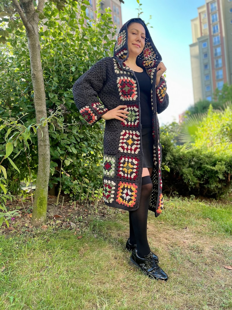 May include: A long, gray crochet cardigan with a hood. The cardigan has colorful granny square patches on the front and sleeves. The model is wearing black tights and black shoes.