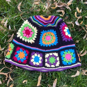May include: A colourful crocheted hat with a black background and a variety of coloured squares. The hat is lying on a bed of green grass with brown leaves.