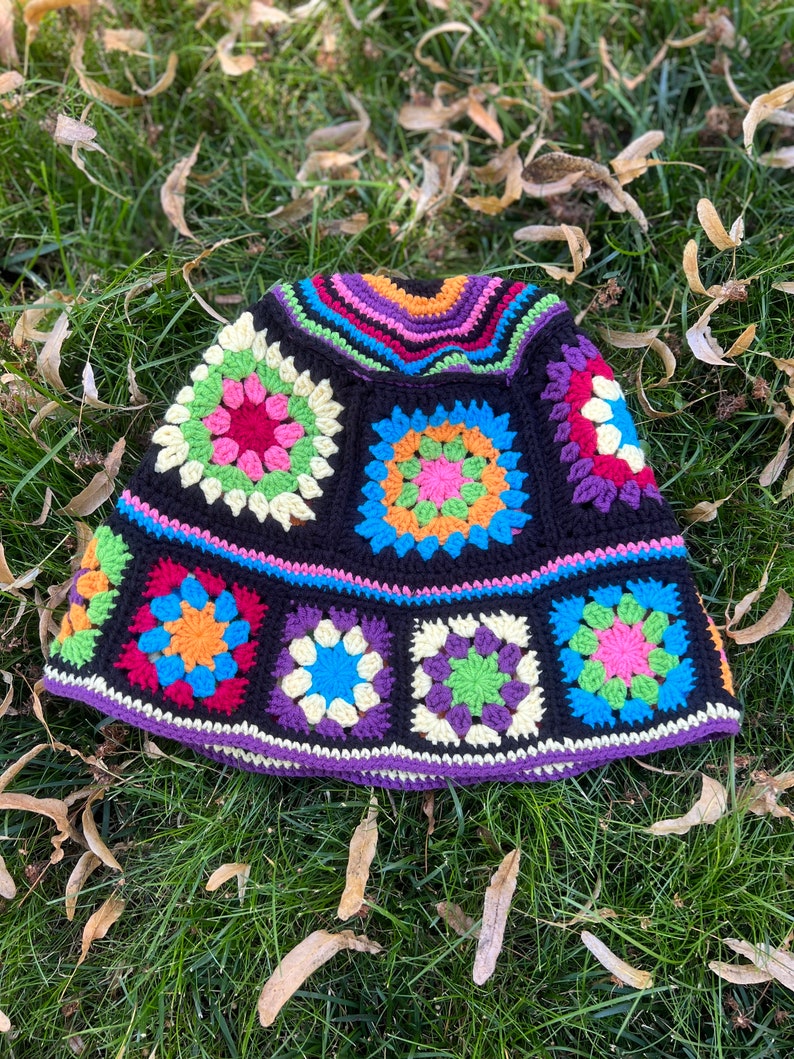 May include: A colourful crocheted hat with a black background and a variety of coloured squares. The hat is lying on a bed of green grass with brown leaves.