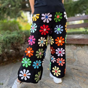 May include: Black crochet trousers with a colourful daisy pattern. The trousers are wide-legged and have a relaxed fit.