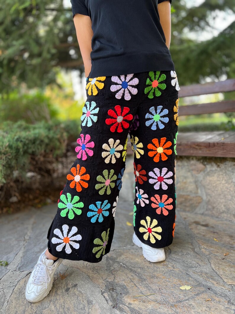 May include: Black crochet trousers with a colourful daisy pattern. The trousers are wide-legged and have a relaxed fit.