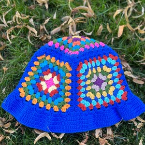 May include: A blue crochet bucket hat with a colourful granny square pattern. The hat is lying on a bed of green grass with brown leaves.
