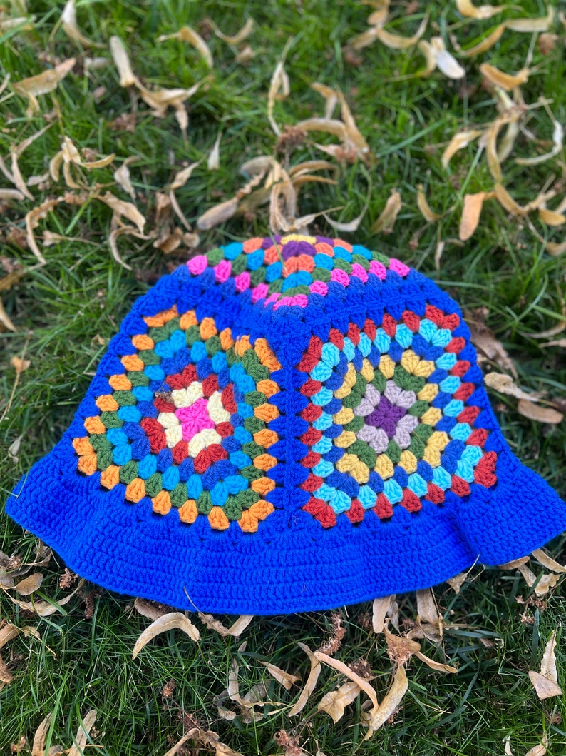 May include: A blue crochet bucket hat with a colourful granny square pattern. The hat is lying on a bed of green grass with brown leaves.