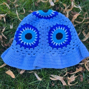 May include: A blue crocheted bucket hat with two large blue evil eye designs. The hat is made with a variety of shades of blue yarn.