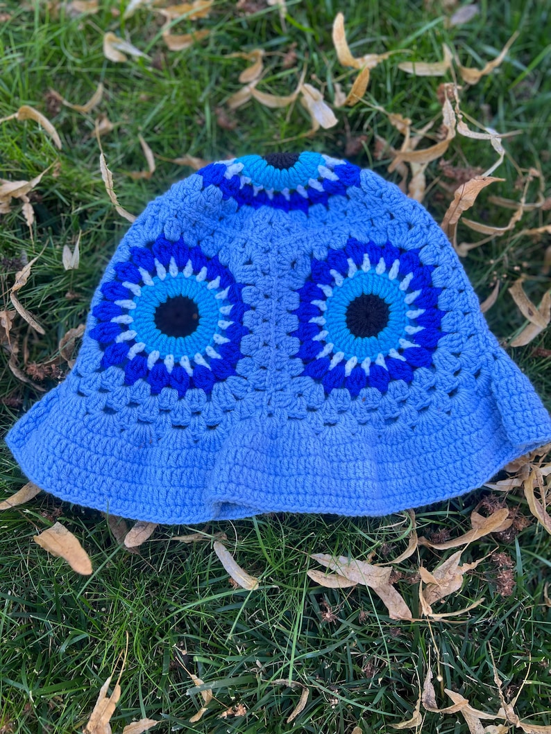 May include: A blue crocheted bucket hat with two large blue evil eye designs. The hat is made with a variety of shades of blue yarn.