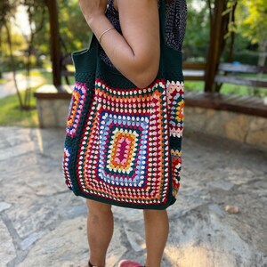 May include: A large green crochet tote bag with a colorful geometric pattern. The bag has a square shape with a large central square in shades of pink, orange, yellow, green, and blue. The bag is made with a thick yarn and has a sturdy construction.