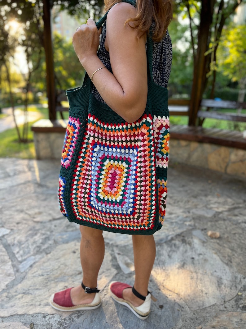May include: A large green crochet tote bag with a colorful geometric pattern. The bag has a square shape with a large central square in shades of pink, orange, yellow, green, and blue. The bag is made with a thick yarn and has a sturdy construction.