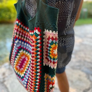May include: A green crochet tote bag with a colorful granny square pattern. The bag has a long strap and is made of thick yarn.