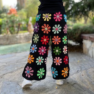 May include: Black crochet trousers with a colourful daisy pattern. The trousers are wide-legged and have a relaxed fit.