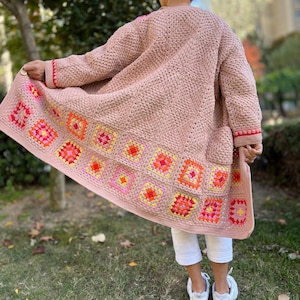 May include: A long, open-front crochet cardigan in a soft pink hue. The bottom edge features a colorful geometric pattern in shades of orange, yellow, and pink. The sleeves and pockets are a solid pink, with red trim. The cardigan is knee-length.