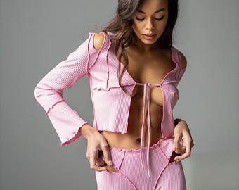 May include: A light pink ribbed knit set. The top has long sleeves, shoulder cutouts, and a tie closure. The set includes matching high-waisted trousers. Decorative stitching is visible on the edges of the top and trousers.