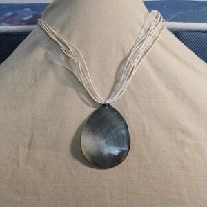 May include: A necklace featuring a large, teardrop-shaped pendant made of iridescent dark gray and white shell material. The pendant hangs from a multi-strand cord necklace made of thin, light cream-colored fabric ribbons that gather at the top.