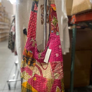 Puede incluir: Un bolso de hombro de tela de patchwork rosa y multicolor con una correa larga. El bolso está hecho de tela acolchada y tiene una variedad de patrones y colores.