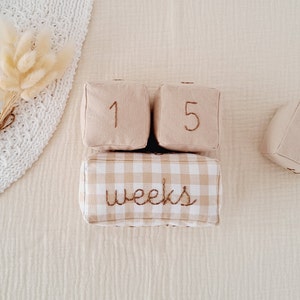 Pode incluir: Blocos de tecido bege com os números 1, 5 e 3, e um bloco vichy com a palavra "semanas" bordada.