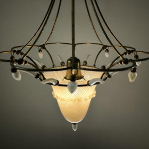 May include: An antique chandelier with a brass frame and a frosted glass shade. The chandelier has a decorative design with white accents and hanging crystal-like ornaments. The light is on, illuminating the intricate details.