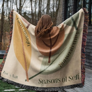 Peut inclure: Une couverture tissée beige avec un motif de feuilles dans les tons marron, vert et jaune. La couverture porte les inscriptions "I am enough" et "I am growing", ainsi que les mots "Seasons of Self" et "Zen & Sass". Elle est bordée de franges.