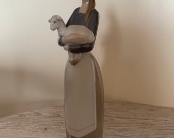 May include: A porcelain figurine of a woman holding a white lamb in her arms. The woman is wearing a brown dress with a white apron and has brown hair. The figurine is standing on a green base.