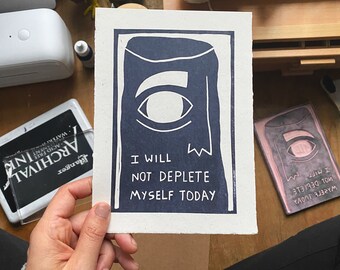 May include: A navy blue and white art print with the text "I WILL NOT DEPLETE MYSELF TODAY" and an eye design. The print is held in a hand, with a stamp and ink pad visible in the background.