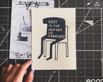 Op de afbeelding: Een zwart-witte illustratie van een persoon die op een stoel zit met de tekst "Rust is de moeder van creativiteit".