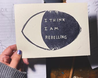 May include: A white card with black ink printed on it. The card has a black eye shape with the text "I THINK I AM REBELLING" inside.