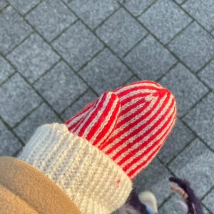Op de afbeelding: Close-up van een rood-witte gestreepte want met een crèmekleurige manchet. De want is gemaakt van een gebreid materiaal. De persoon draagt een bruine jas. De achtergrond is een grijze bakstenen loopbrug.