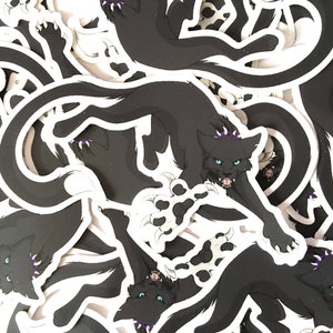 May include: A stack of black and white stickers featuring a cartoon black cat with purple accents and a crown. The cat is depicted with sharp claws and a fierce expression.