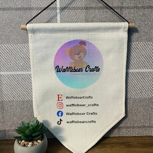 May include: A white canvas banner with a brown wooden dowel at the top and bottom. The banner features a colourful circle with a cartoon bear and the text "Wafflebear Crafts" in black. Below the circle, there are social media icons and usernames for Etsy, Instagram, Facebook, and TikTok.