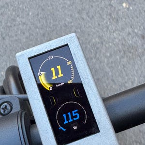 May include: Close-up of an electric scooter's digital display. The screen shows a speed of 11 mph and a power output of 115 W. The display is set in a gray frame, with a black background and yellow and blue digital readouts.