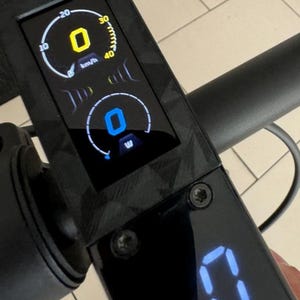 Op de afbeelding: Een zwart dashboard van een elektrische scooter met een digitaal display dat een snelheid van 0 km/h, een batterijniveau van 0% en een blauw digitaal display met 0 laat zien.