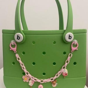 May include: A green tote bag with a chain of pink charms. The bag has two handles and the letter "b" on the front. The charms include a flower, ice cream, and a bear.