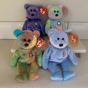 May include: Four colorful plush teddy bears with attached tags. The bears are in shades of purple, blue, green, and peach. Each bear has a different design on its chest. The bears are sitting on a white surface.