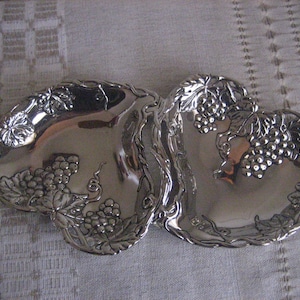 May include: A silver heart-shaped dish with a grapevine design. The dish is made of metal and has a shiny finish.