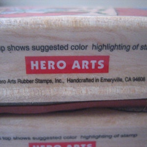 May include: A wooden stamp block with the text "Label top shows suggested colour highlighting of stamp" and "HERO ARTS" in red letters. The bottom of the block has the text "1996 Hero Arts Rubber Stamps, Inc., Handcrafted in Emeryville, CA 94608 USA".