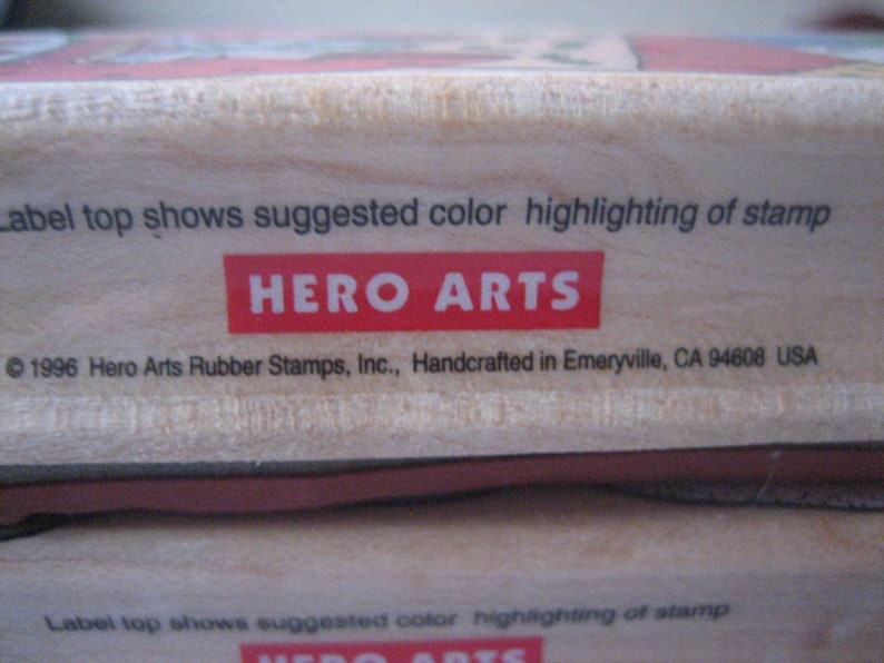 May include: A wooden stamp block with the text "Label top shows suggested colour highlighting of stamp" and "HERO ARTS" in red letters. The bottom of the block has the text "1996 Hero Arts Rubber Stamps, Inc., Handcrafted in Emeryville, CA 94608 USA".