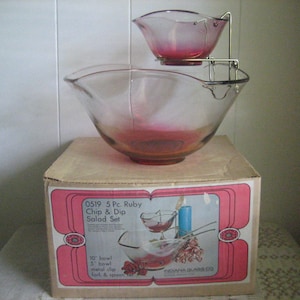May include: A vintage ruby red glass chip and dip salad set with a metal stand. The set includes a 10-inch bowl, a 5-inch bowl, a metal clip, a fork, and a spoon. The box is labeled "0519 5 Pc. Ruby Chip & Dip Salad Set" and "INDIANA GLASS CO."