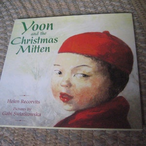 May include: A book cover with a red and white illustration of a child wearing a red hat. The title of the book is "Yoon and the Christmas Mitten". The author is Helen Recorvits and the illustrator is Gabi Swiatkowska.