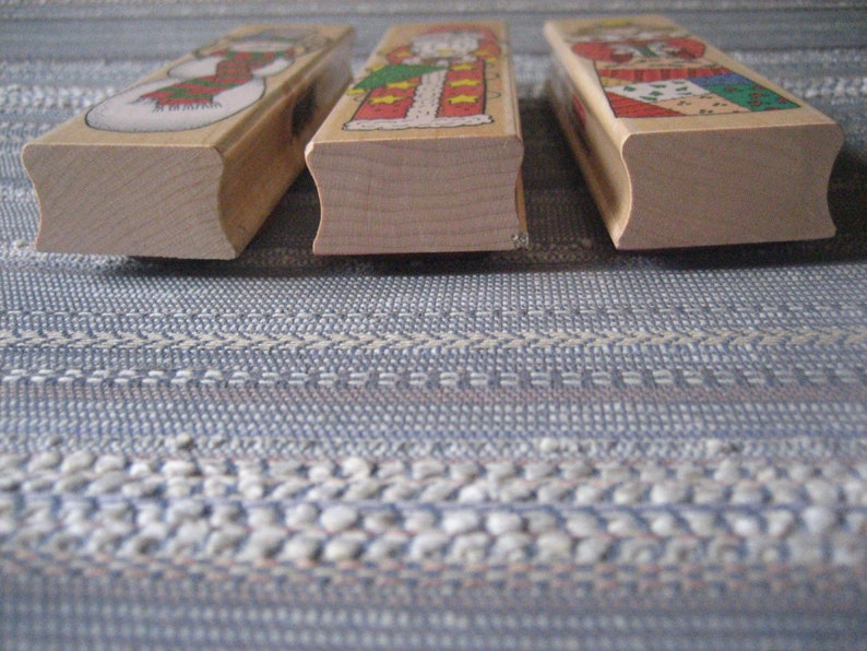 May include: Three wooden stamps featuring Christmas-themed illustrations. The stamps are rectangular with slightly rounded edges. The illustrations depict a snowman, Santa Claus, and a woman in a red dress.