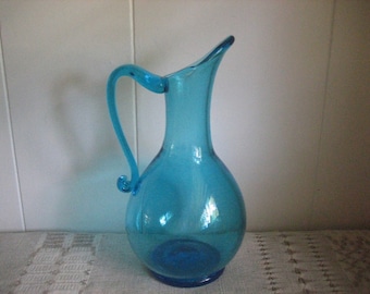 May include: A bright blue glass pitcher with a curved handle. The pitcher has a rounded body and a narrow spout.