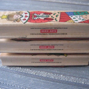 May include: Three wooden mounted rubber stamps with a Christmas theme. The top stamp features a girl in a red and green dress with a star on her head. The middle stamp features a red and green patchwork design. The bottom stamp features a green and red Christmas tree design. All three stamps have the text "HERO ARTS" on the bottom.