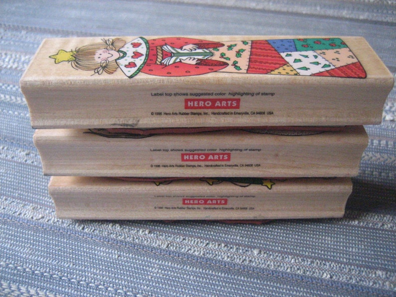 May include: Three wooden mounted rubber stamps with a Christmas theme. The top stamp features a girl in a red and green dress with a star on her head. The middle stamp features a red and green patchwork design. The bottom stamp features a green and red Christmas tree design. All three stamps have the text "HERO ARTS" on the bottom.